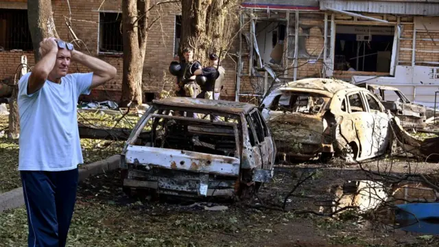 A man holds his hands atop his head as he stands near two burned-out cars