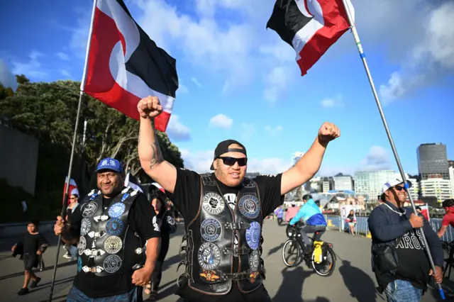 Two men in leather vests covered in badges and patches. One of the men is holding a flag and the other has two fists raised