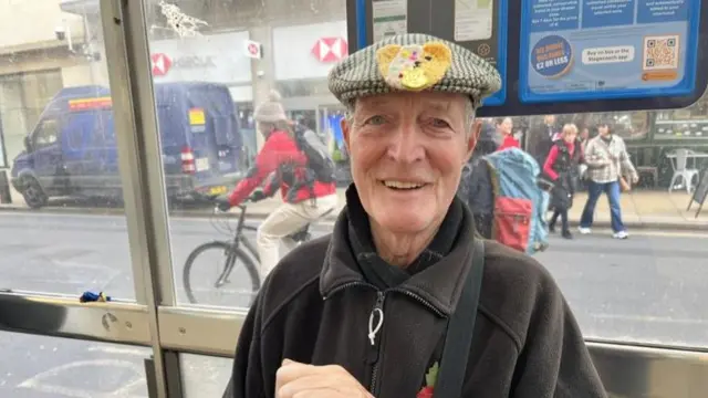 Trevor Minchley sitting in a bus stop in Cambridge city centre, with the street scene visible through the glass behind him