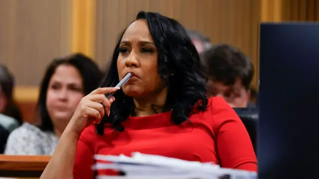 Fani Willis, Fulton County District Attorney, is seated behind a table in a courtroom. She wears a red dress and has contemplatively placed a pen against her top lip