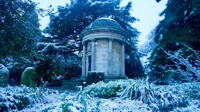 A brick structure among snowy bushes and trees. The structure has pillars around the side and is circular in nature