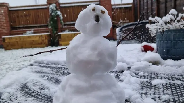 A snowman in a garden. The garden has a dusting of snow on raised beds and a fence. The snowman has twig arms and stones for eyes and a nose