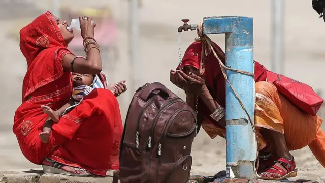 Two women drink water and splash their face from a public tap on a street in Prayagraj, India, on 10 June. They both wear red, with one holding a baby and a backpack on the ground next to them.