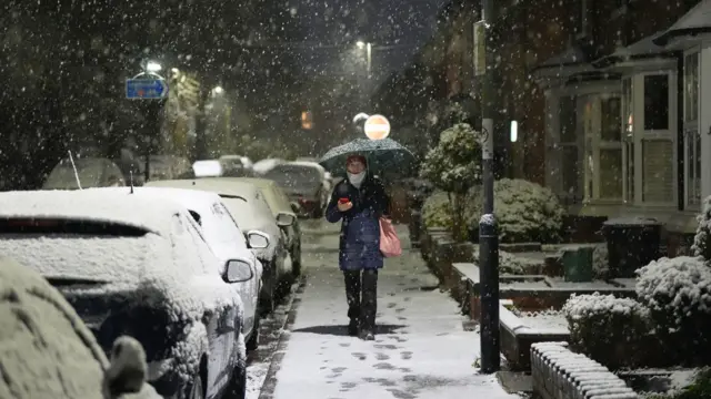 A person walks with an umbrella along a snowy street in Warwick. Alongside, cars are covered in snow