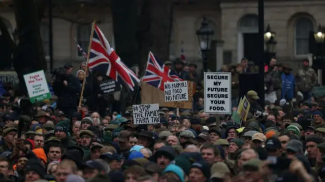 A crowd of people, you can only see their heads, as they hold up signs in protest to the farming inheritance announcement