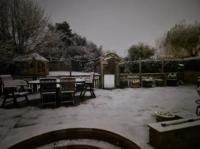 A snow-covered garden with chairs, a table, a fence and an archway surrounded by a hedge