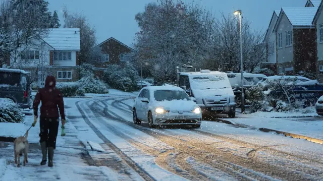 A person walks a dog on a snowy street while a car drives past with snow on its bonnet. Around them, snow dusts on houses and pavements