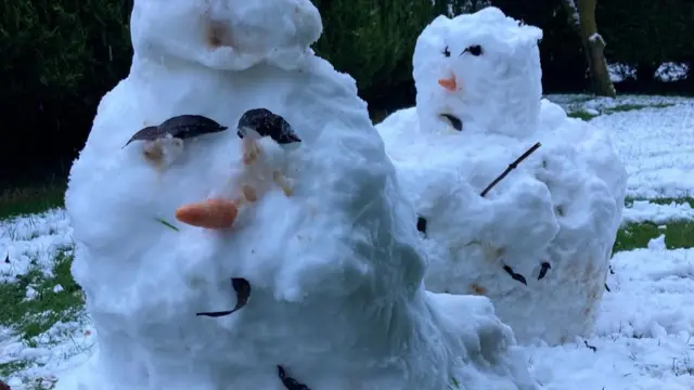 Two snowmen in a garden. They both have carrots for noses, leaves for eyes and a mouth. The garden has a dusting of snow with green patches of grass showing in places