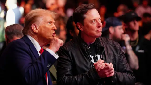 Donald Trump clenches his fist while standing next to Elon Musk who has his hands clasped together