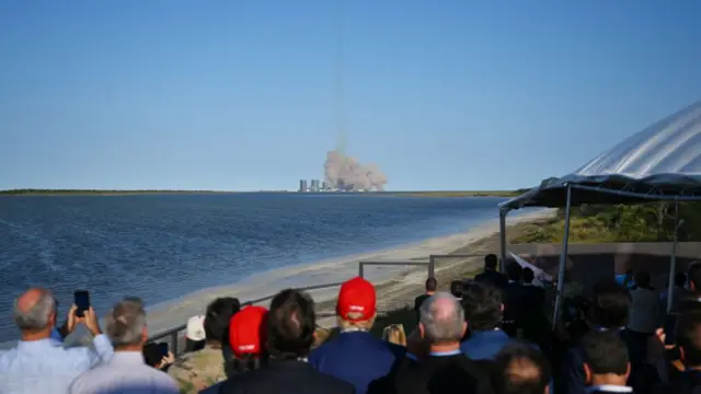 The back of a crowd of peoples heads, with Trump in the middle in a bright red cap, as the rocket launches across a body of water in front of them