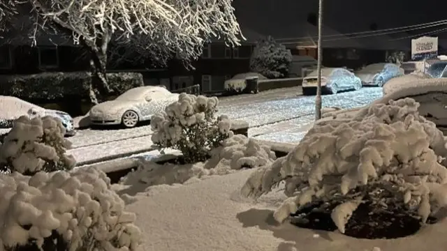 Snow in a street in Leek with cars and gardens covered