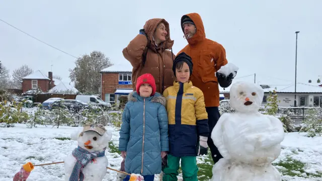 A family of four wearing coats in the snow, with a snowman on either side of them and the dad is holding a large snowball in his hand and looking at his wife