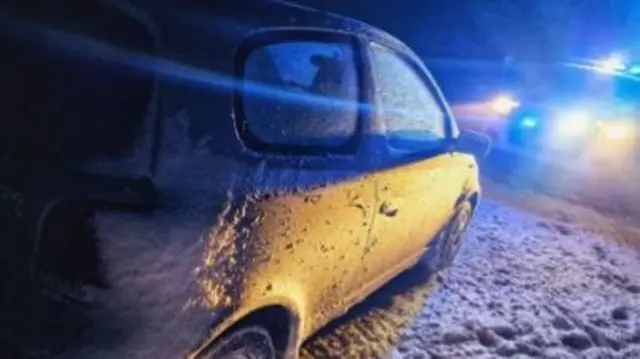 Silver car covered in snow on a road with a police car ahead of it with its lights on