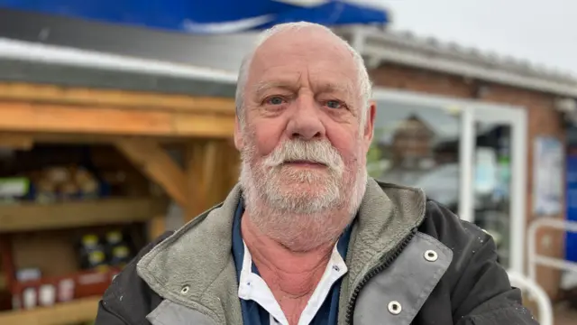 A man with white hair and beard in a brown fleece lined jacket, standing in front of a shop