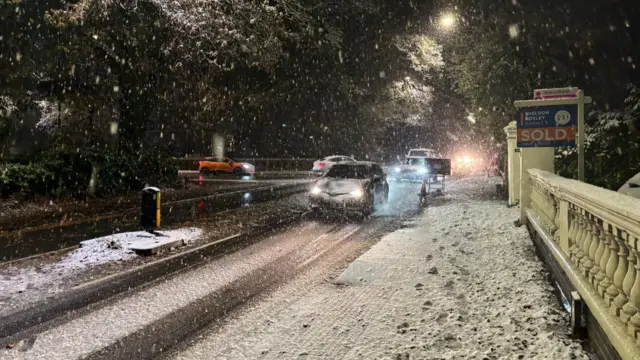 Several cars driving through snow in Leamington, there is snow on the pavement too and footprints can bee seen there