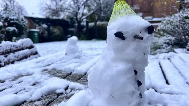 A small snowman in a garden covered in snow.  An upturned green shuttlecock has been used as his hat while stones make his eyes and a twig his nose