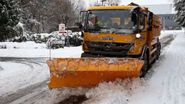 A gritting lorry on a snow-covered road with a yellow snow plough at the front