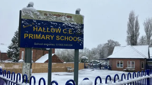 A school covered in snow with a blue sign in the foreground reading Hallow CE Primary School