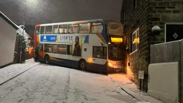 A double decker bus blocks a steep, snow-covered road after skidding and coming to rest against the side of a house.