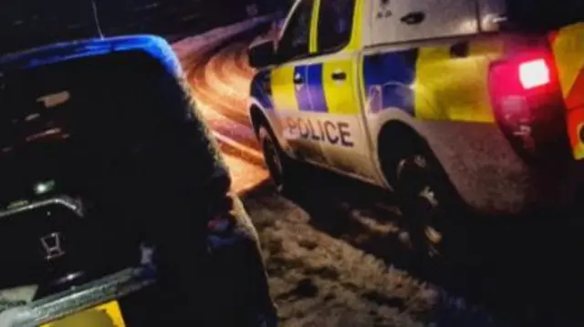 Black car abandoned on a road with snow and police car parked alongside it