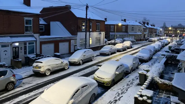 Cars covered with snow on a road near houses in Bearwood, Smethwick, with snow covering the pavement and tyre tracks showing on the road from some cars making the journey