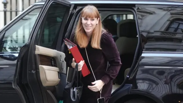 Angela Rayner getting out of a black Range Rover, holding a red folder and a black handbag with her phone dangling around her on a shoulder strap