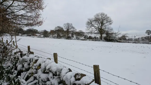 A stone wall in Ipstones covered in stone with a fence of barbed wire behind it which is in front of a snow-covered field leading to some trees in the distance