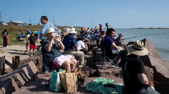 A crowd of people sitting by the water with binoculars and smart phones. One man fishes