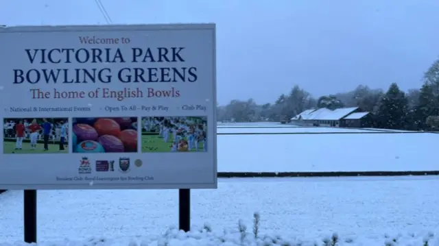 A sign which says Victoria Park Bowling greens, with snow on the grass behind