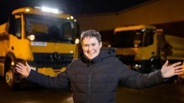 Councillor Grace Baynham smiling with her arms stretched out in front of Stockport Council's gritters