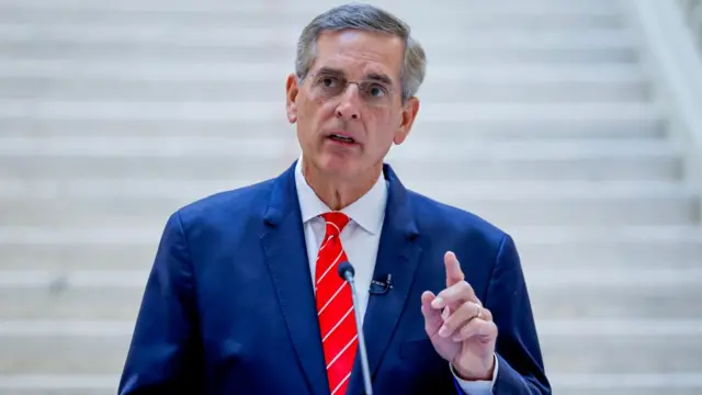 Brad Raffensperger speaks at a podium in a blue suit and red tie