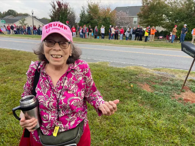 A woman, called Becky, smiles at the camera. She wears a pink cap featuring the words "Trump 2020", a pink paisley shirt, and carries a coffee cup. A queue of Trump rally-attendees are seen behind her.