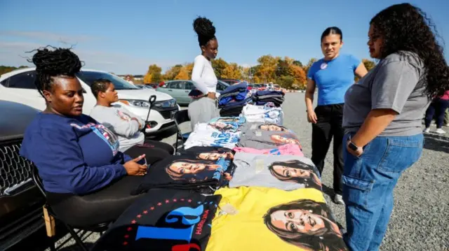 Women sitting and standing around tables covered with black, yellow, pink, white and grey T-shirts, all with an image of Kamala Harris on them