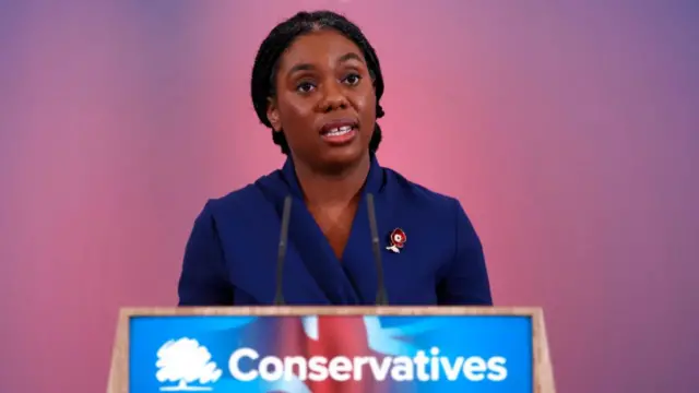 Kemi Badenoch mid-shot as she stands behind a podium marked Conservatives. She's in a blue dress with a poppy enamel pin on her left lapel and her hair pulled back in a low bun