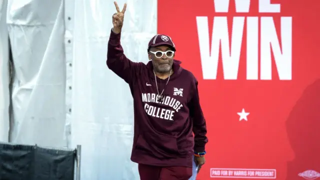 Spike Lee, wearing a maroon matching tracksuit, holds up a peace sign at a Harris rally in Clarkston, Georgia