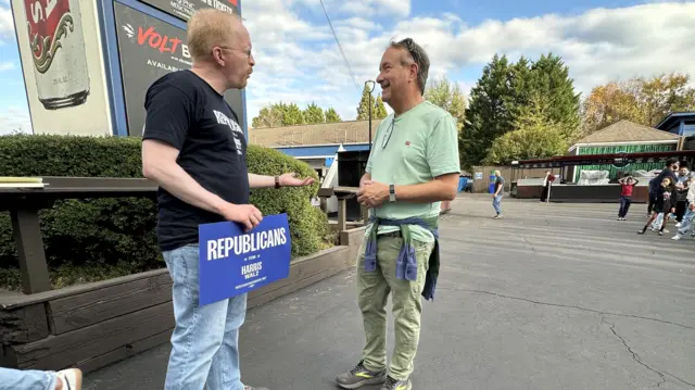 Two men stand and speak to one another holding Republicans for Harris/Walz signs