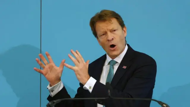 Richard Tice speaking on stage in black suit, turquoise patterned tie and white shirt. He has both hands raised and angled to his right