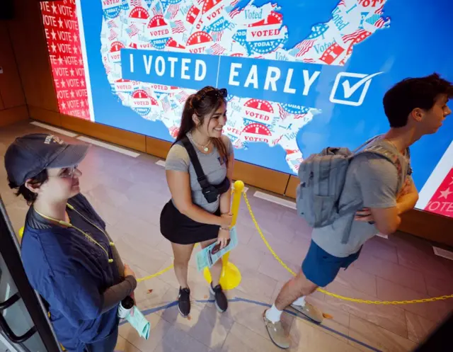 Three people wait in line with a large graphic in the background titled I Voted Early with a map of the US