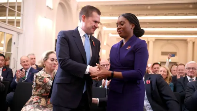 Robert Jenrick in blue suit, light blue tie and white blouse shakes hands with Kemi Badenoch, who's wearing a Conservartive blue dress. They're both wearing poppy pins