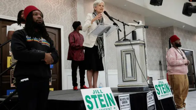 Jill Stein speaks at a podium with signs that say Jill Stein for president. She is wearing a keffiyeh scarf