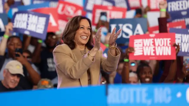 Kamala harris stands in front of a clapping crowd, and wears a tan suit. She is also clapping and smiling.