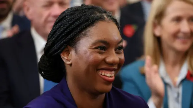 Close-up shot of Kemi Badenoch smiling. She's in a Conservative blue dress, her hair pulled back in a low bun. Background is blurred crowd