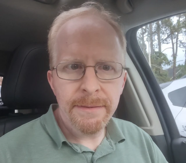 A selfie of Michael Tucker sitting inside a vehicle. He has glasses, strawberry-blonde hair and a beard.