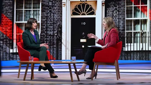 Racehel Reeves in forest green pant suit with bluer shirt and black heels sitting down. Sitting in front of her is Laura Kuenssberg in a hot pink jacket, black pencil trousers and heels. She has her right had raised towards Reeves. A close-up photo of Number 10's front door is in the background