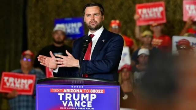 JD Vance stands at a podium with his name and Donald Trump's on it and gestures to the crowd.