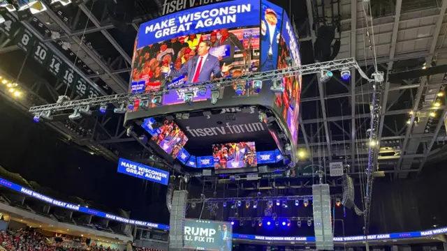 Donald Trump rally in a large stadium shows a man speaking on a jumbotron, with the words: Make Wisconsin Great Again.