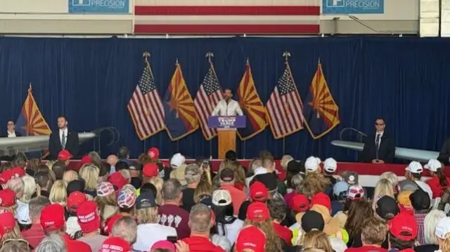 Donald Trump Jr speaks at a podium, members of the crowd wear red MAGA hats