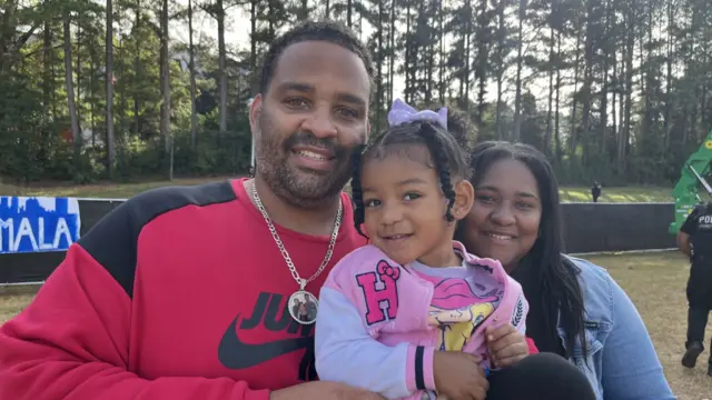 A man wearing a red sweatshirt with his wife, holding their young daughter in the middle, at a Kamala Harris rally in Atlanta, Georgia