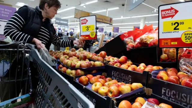 A woman shops at the grocery store and holds up apples to compare prices.