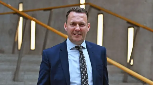 Russell Findlay walks down steps of Scottish Parliament. He's in a dark blue suit with light blue shirt and patterned blue tie